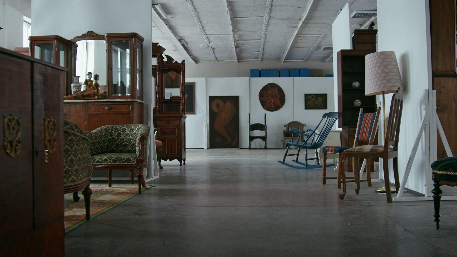 A long antiques showroom: carved wooden vitrine cabinets, a green damask armchair, and a blue rocking chair on a polished concrete floor